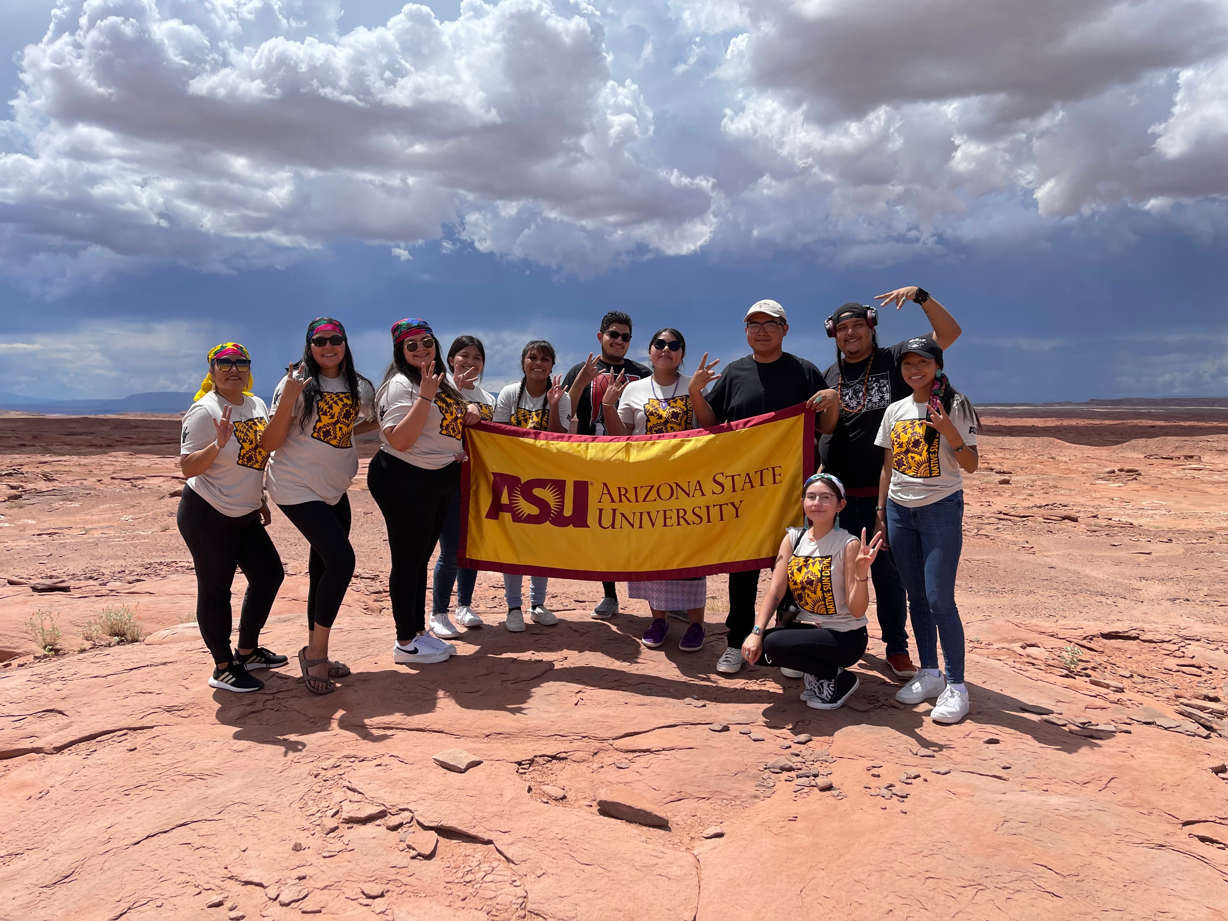 Group of students outside in desert wearing "native sun devil" shirts and holding ASU banner