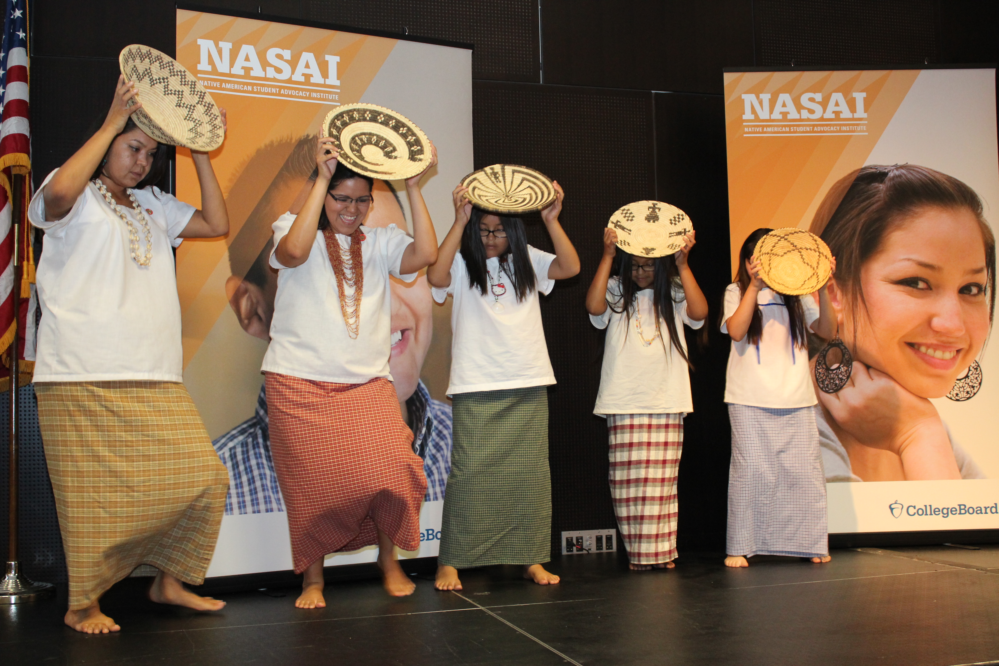 Five people dressed in white shirt and long skirts holding woven basket above their heads while dancing